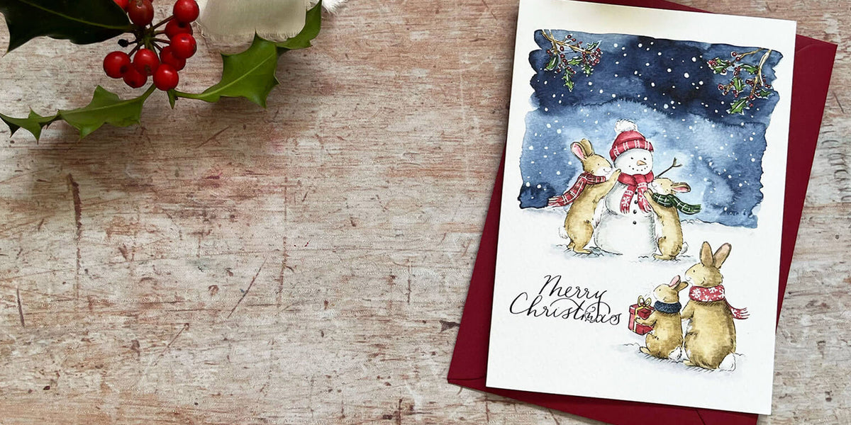 A Christmas greeting card featuring watercolor illustrations of rabbits and a snowman in a snowy night scene, placed on a red envelope over a rustic wooden surface with holly leaves and red berries in the corner.